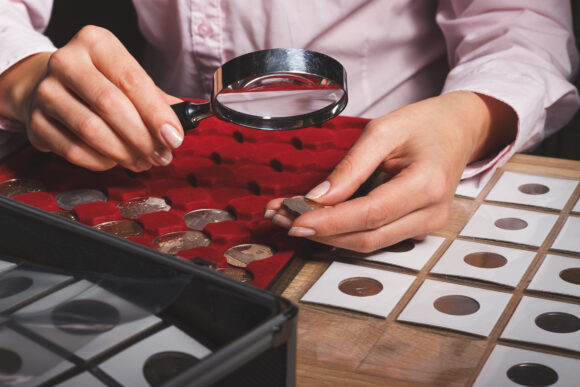 Box with collectible coins in the cells and a hand with coin through the magnifying glass, soft focus background